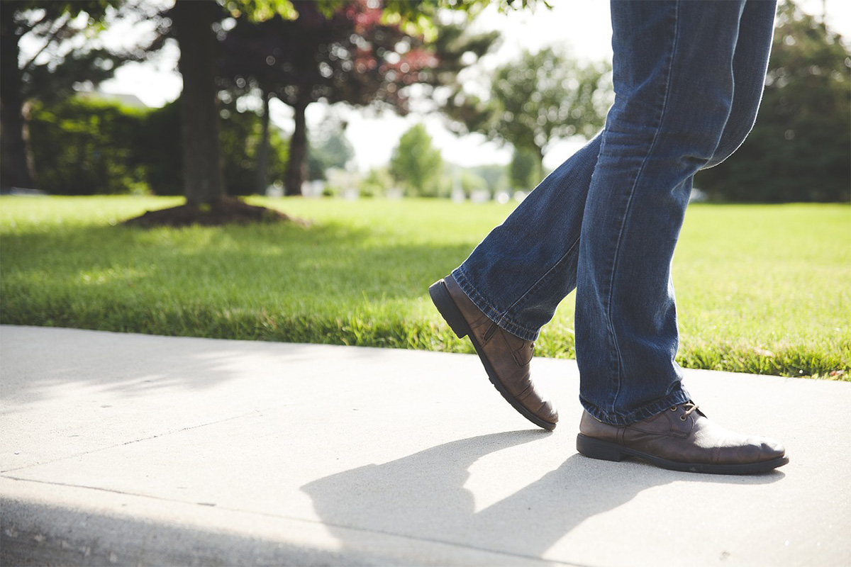 Person wearing blue jeans and brown shoes walking on a concrete sidewalk in a park-like setting with grass and trees in the background, reminiscent of the scenic outdoor spaces found in Sterling Ranch CO within the Denver metro area.