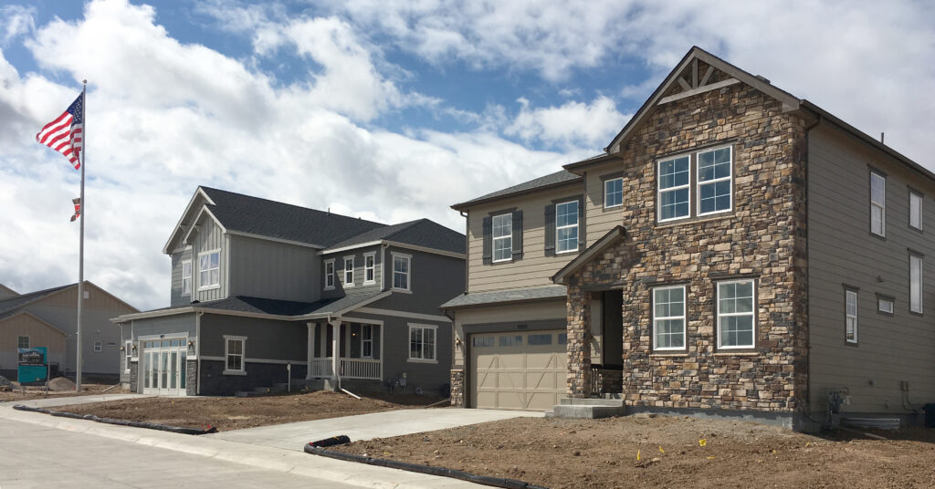 Two newly built, two-story suburban houses with stone and siding exteriors are shown on a cloudy day in Sterling Ranch CO, with an American flag on a pole and unfinished landscaping in front.