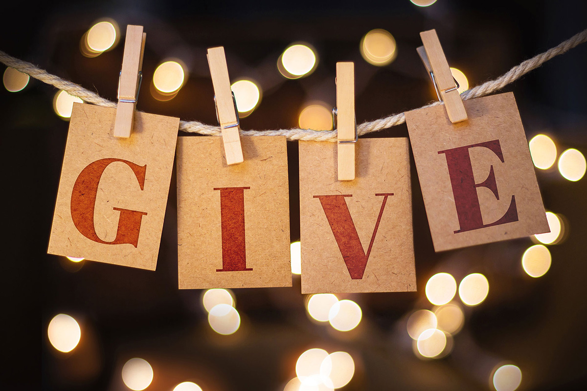 Four cards spelling "GIVE" hang from a string with clothespins, set against a backdrop of blurred warm lights—a heartfelt scene reminiscent of the warmth and generosity found in Sterling Ranch, CO.
