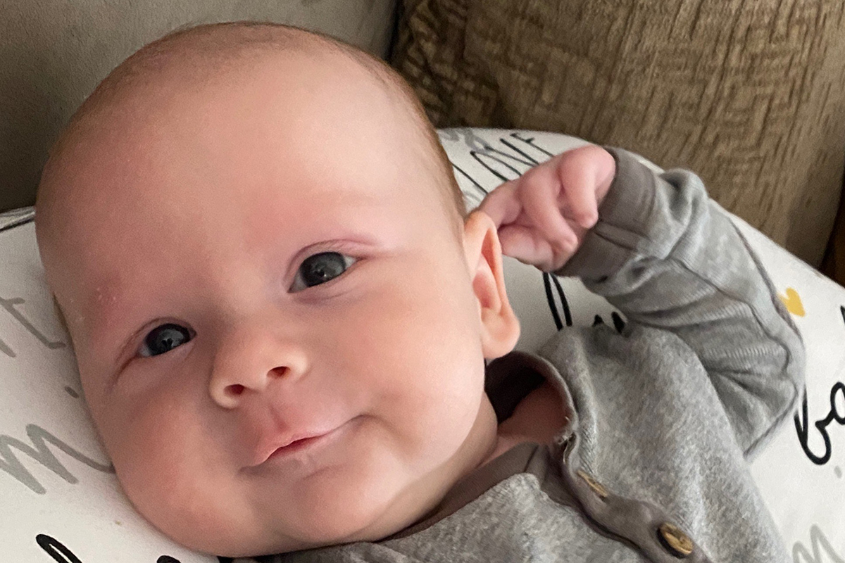 A baby wearing a gray outfit lies on a patterned pillow in a cozy Sterling Ranch Colorado home, looking at the camera with a relaxed expression and one hand resting near their head.