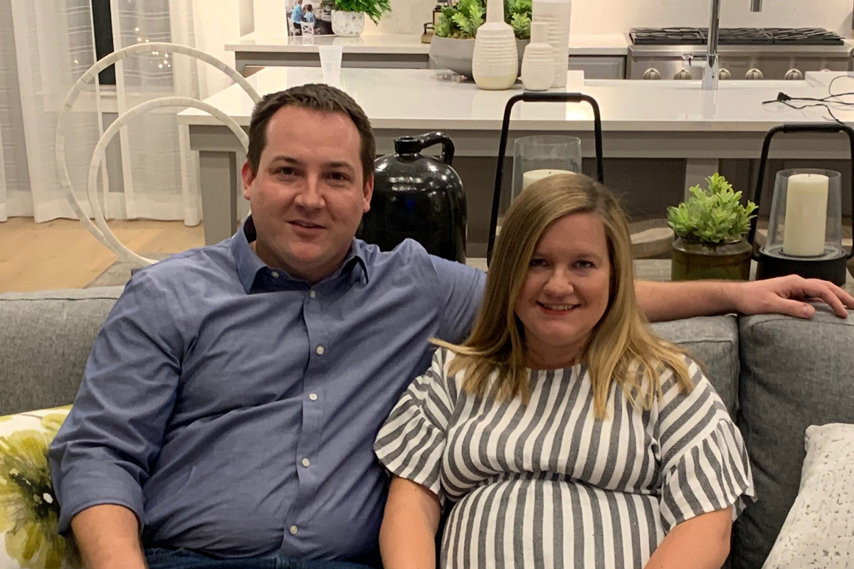 A man and a pregnant woman sit together on a gray couch in a modern living room, smiling at the camera in their beautiful Sterling Ranch home just southwest of Denver.