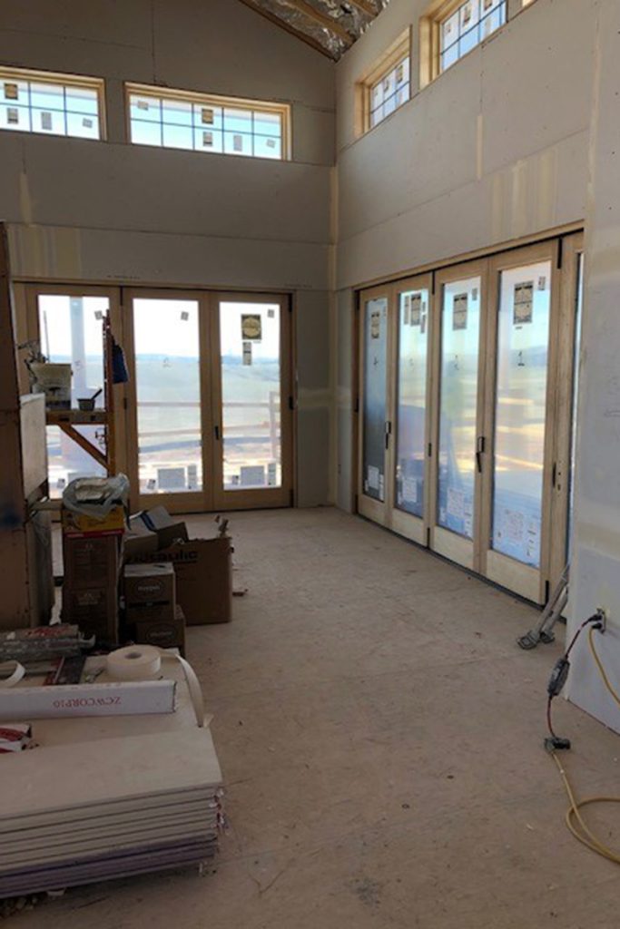 Partially finished room with large floor-to-ceiling windows and sliding doors, exposed drywall, construction materials, and tools scattered on the floor.