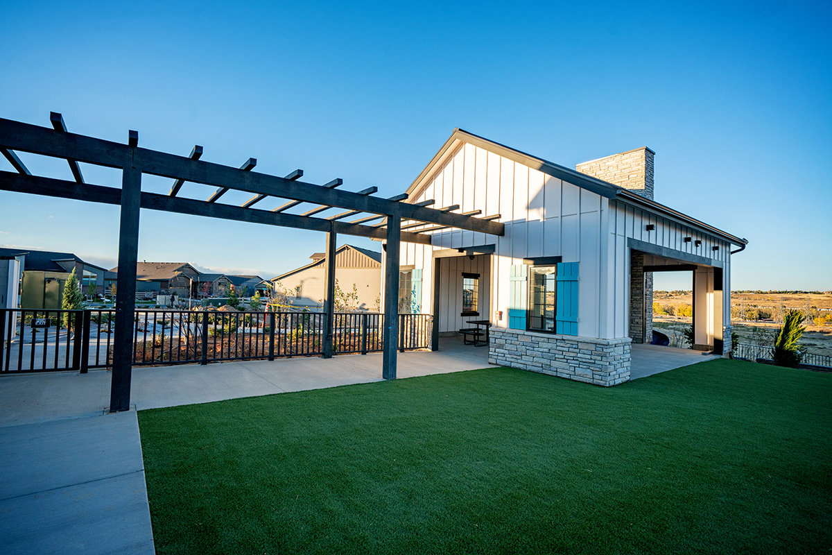 Modern house with white siding, stone accents, and a covered patio in the desirable Sterling Ranch CO community. A pergola and black fence border a landscaped yard with artificial turf under a clear sky.