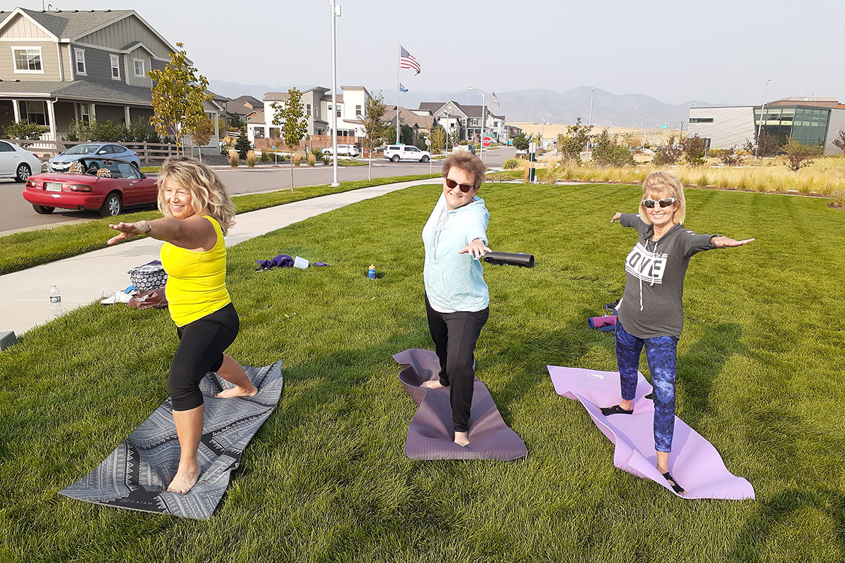 Three women practice yoga outdoors on mats in a grassy area, performing the warrior pose, with houses and mountains visible in the background. This serene scene takes place in Sterling Ranch Colorado, southwest of Denver.