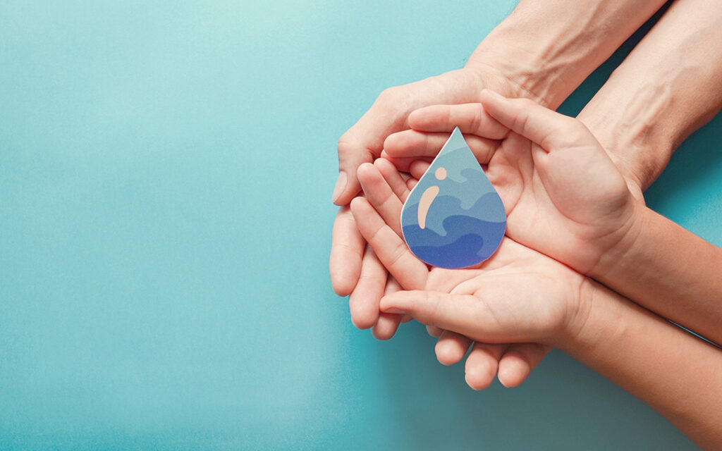 Two adult hands and one child's hands cradle a paper cutout of a water droplet with wave designs, set against a light blue background—reflecting the community spirit and commitment to sustainability found in Sterling Ranch Colorado, near the Denver metro area.