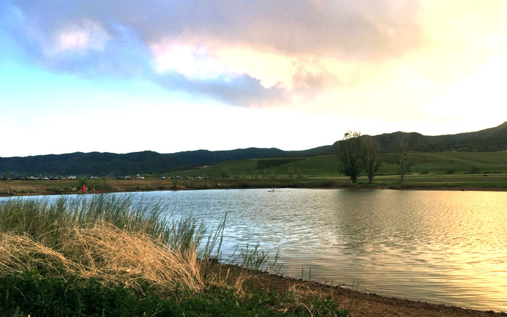 A calm lake bordered by grass and shrubs, with rolling hills and a partly cloudy sky in the background during sunset, evokes the peaceful landscapes southwest of Denver in Littleton, CO.