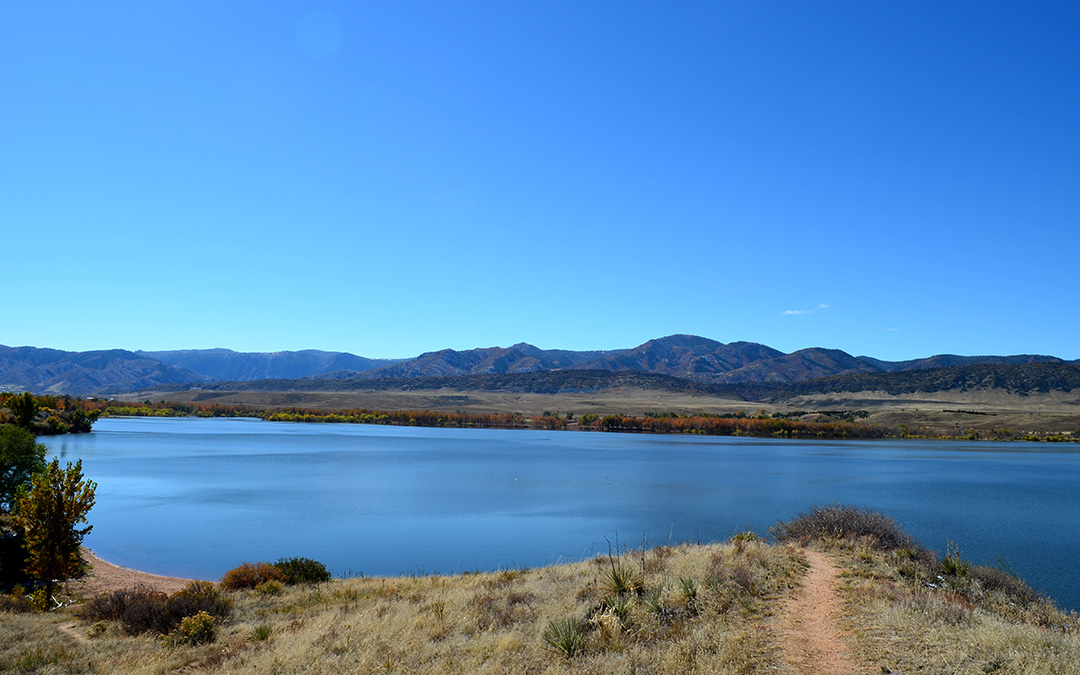 A calm lake with a dirt path in the foreground, surrounded by dry grasslands and trees, set against the distant mountains under a clear blue sky—capturing the serene natural beauty of Sterling Ranch Colorado.