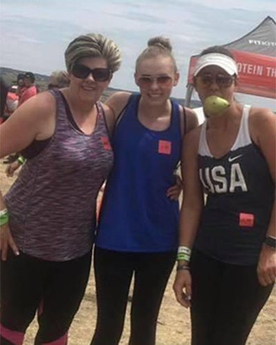 Three women pose outdoors at an event in Sterling Ranch CO; one wears a “USA” tank top and holds a green apple in her mouth. All wear sports attire and sunglasses, with event wristbands and numbered stickers.