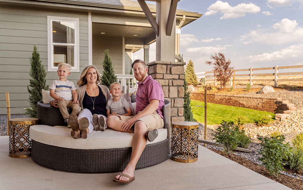 A family of four sits together on a round outdoor sofa on a patio in Sterling Ranch CO, smiling at the camera, with a house and landscaped yard in the background.