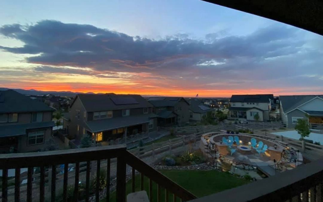 View from a balcony overlooking suburban houses in Sterling Ranch CO, a circular patio with chairs, and a colorful sunset sky with scattered clouds.
