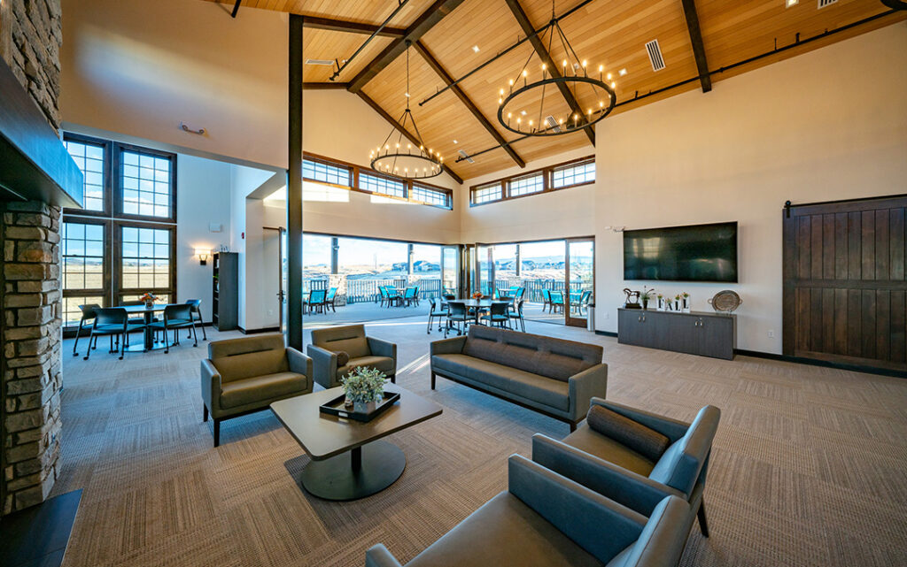 Spacious lounge with modern chairs, large windows, circular chandeliers, a fireplace, TV, and outdoor seating area visible through glass doors—offering the perfect retreat in Sterling Ranch Colorado, southwest of Denver.