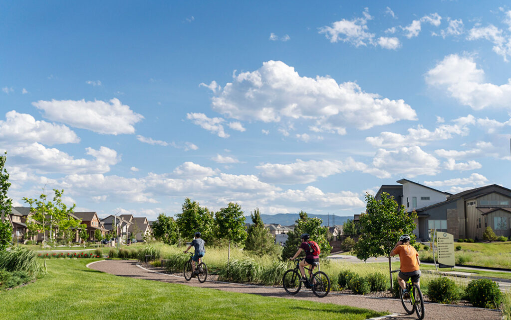 Three cyclists ride on a paved path through a suburban park with grass, trees, and modern houses under a partly cloudy sky in Sterling Ranch, Colorado, southwest of Denver.