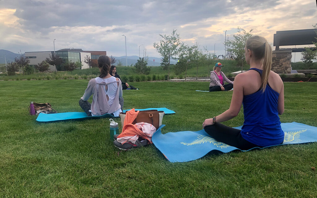 Several people sit on yoga mats on a grassy lawn in Sterling Ranch CO, facing away, with outdoor buildings and a cloudy sky in the background.