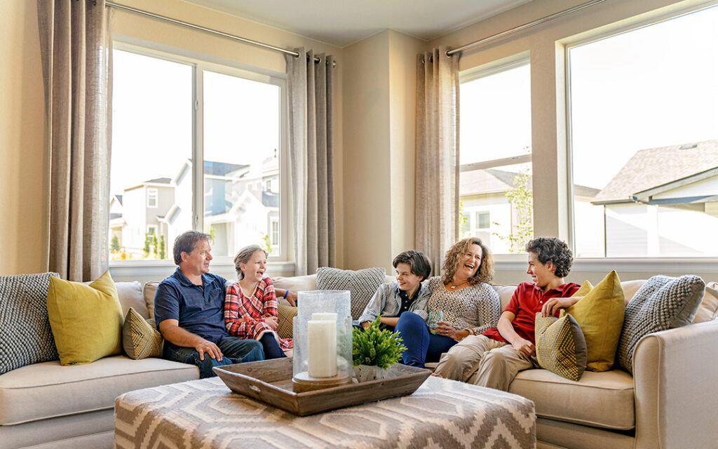 Five people sit on a beige sectional sofa in a bright Sterling Ranch living room, smiling and talking. Large windows reveal houses outside in Littleton, CO. A tray with candles and greenery is on the coffee table.