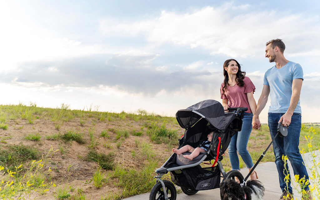 A man and woman walk outside holding hands, pushing a stroller with a child and walking a dog on a paved path beside a grassy area under a partly cloudy sky in Sterling Ranch CO.