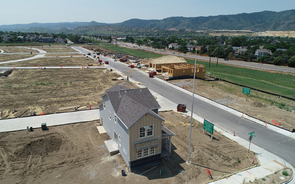 Aerial view of Sterling Ranch Colorado, a residential housing development under construction, featuring a finished house, framed houses, roads, and open lots, all set against a backdrop of hills and greenery.