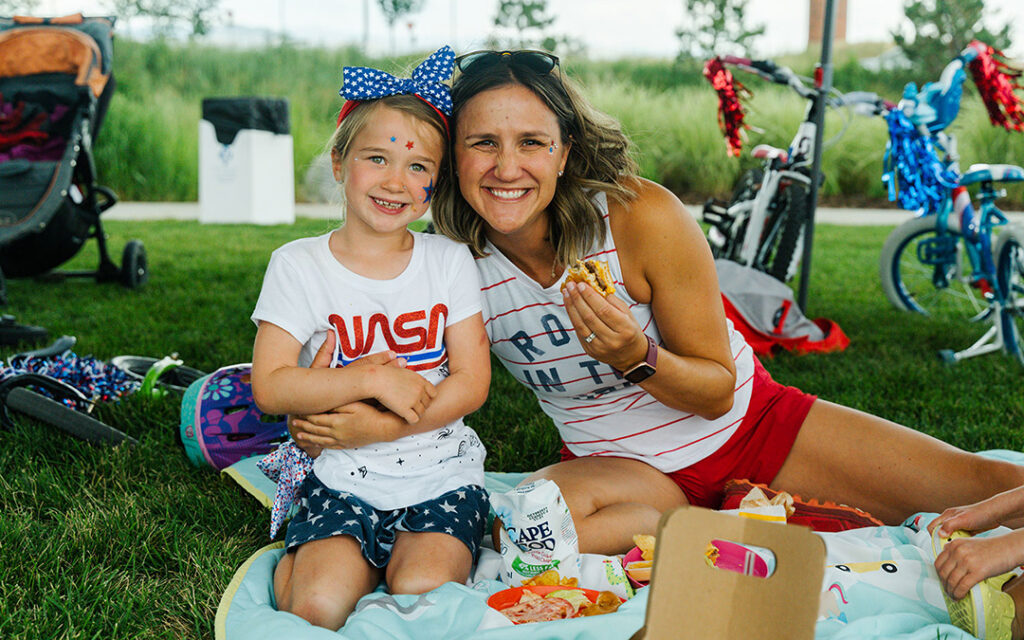 A woman and a young girl sit on a picnic blanket outdoors in Sterling Ranch, southwest of Denver, smiling at the camera with snacks and drinks in front of them.