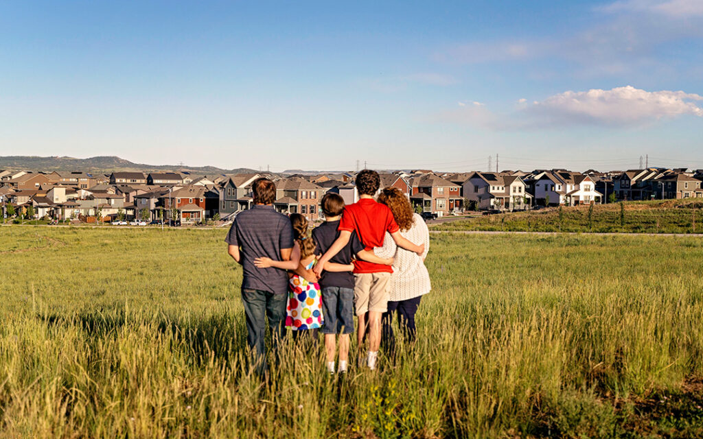 Five people stand together in a grassy field, facing away and looking toward the Sterling Ranch CO neighborhood under a blue sky with scattered clouds, capturing the welcoming spirit of the Denver metro area.