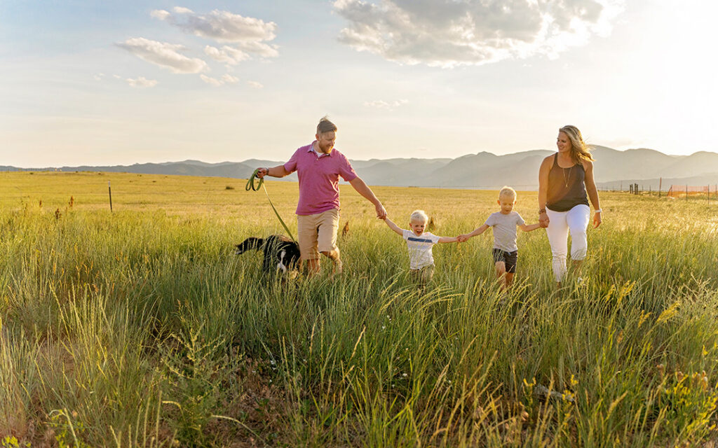 Two adults and two children walk hand in hand through a grassy field with a dog on a leash, mountains and clouds visible in the background of Sterling Ranch Colorado, just southwest of Denver.
