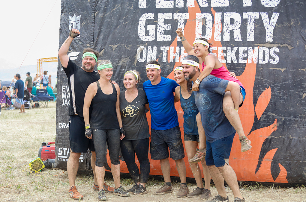 Seven people in athletic gear, some muddy, pose in front of a large event sign at Sterling Ranch Colorado. One person is being carried piggyback, and another raises a fist in celebration.