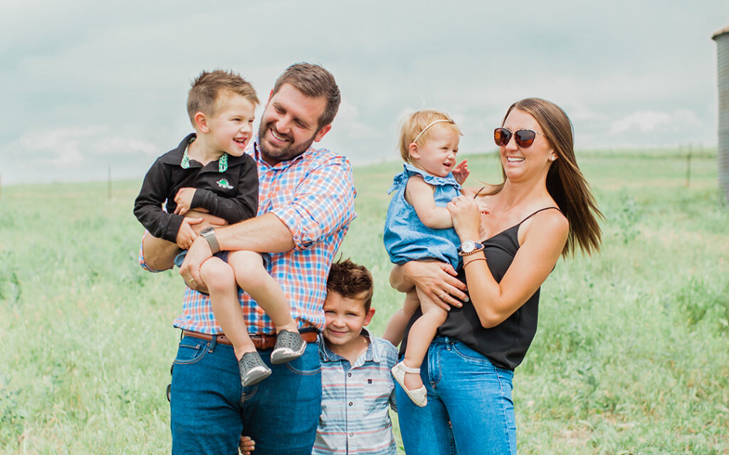 A family of five stands in a grassy field at Sterling Ranch Colorado, southwest of Denver. Two adults hold a young boy and a toddler girl; another young boy stands between them. All are smiling and dressed casually.