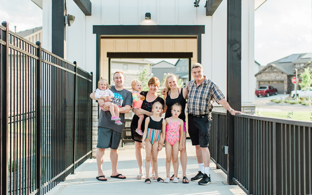 Seven people, including two men, three women, and two young girls, stand together on a sunlit walkway in Sterling Ranch CO, smiling at the camera. One man is holding a baby. Houses are visible in the background, capturing the welcoming atmosphere of this Denver metro community.