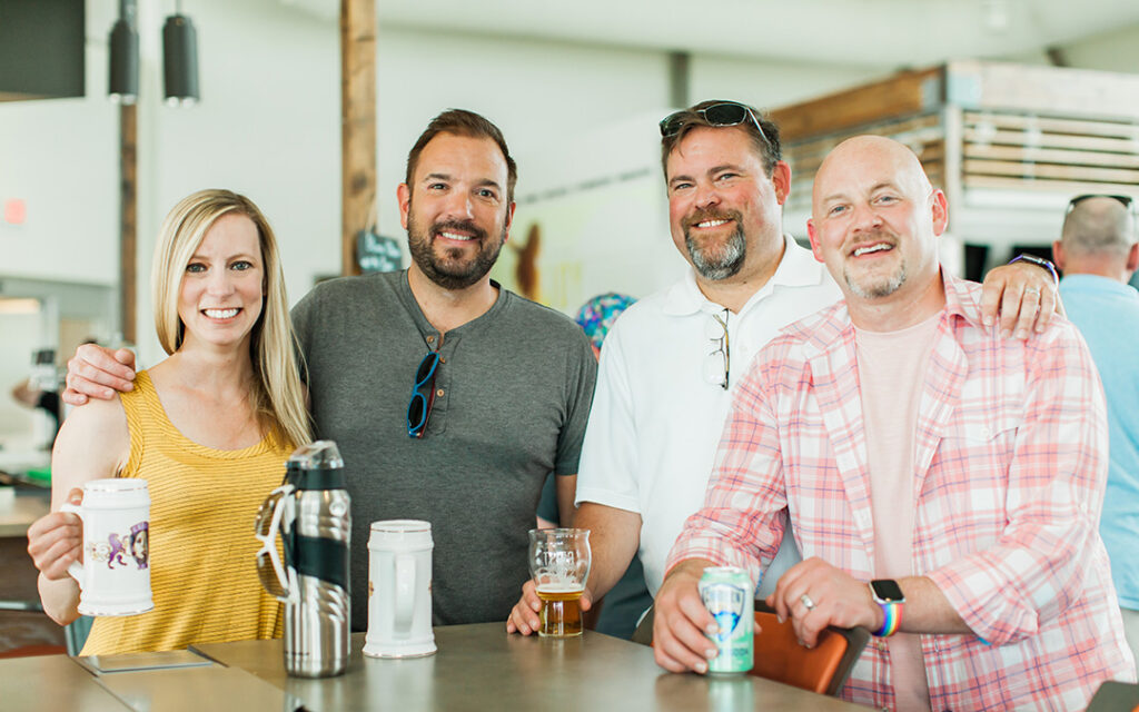 Four adults stand together at a counter in a casual and relaxed setting, smiling and holding drinks. Cups and mugs are arranged on the counter in front of them as they enjoy each other's company. The scene could easily take place in a welcoming home southwest of Denver, perhaps in the inviting community of Sterling Ranch within the Denver metro area.