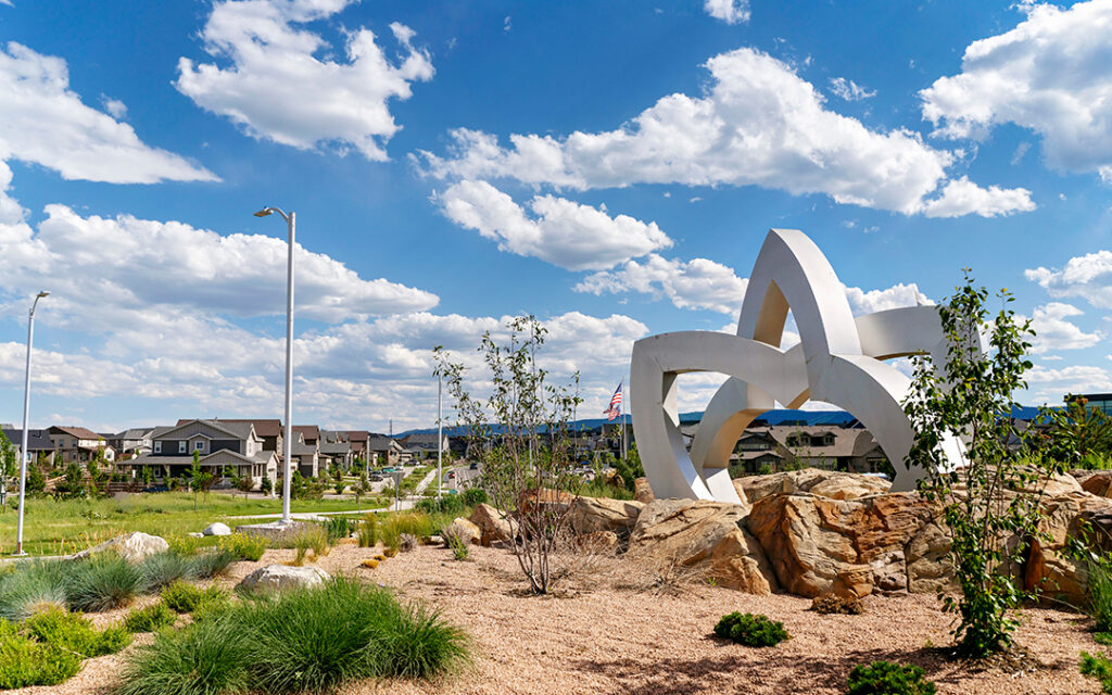 White abstract sculpture stands on rocks in a landscaped area with grass, shrubs, and houses in the background under a blue sky with scattered clouds at Sterling Ranch Colorado, southwest of Denver.