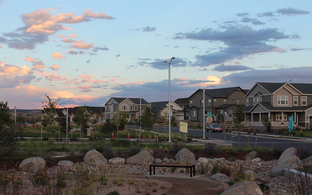 A suburban neighborhood street at sunset in Sterling Ranch CO, with modern houses, a bench, rocks, and landscaping in the foreground under a partly cloudy sky. This welcoming community is located southwest of Denver.