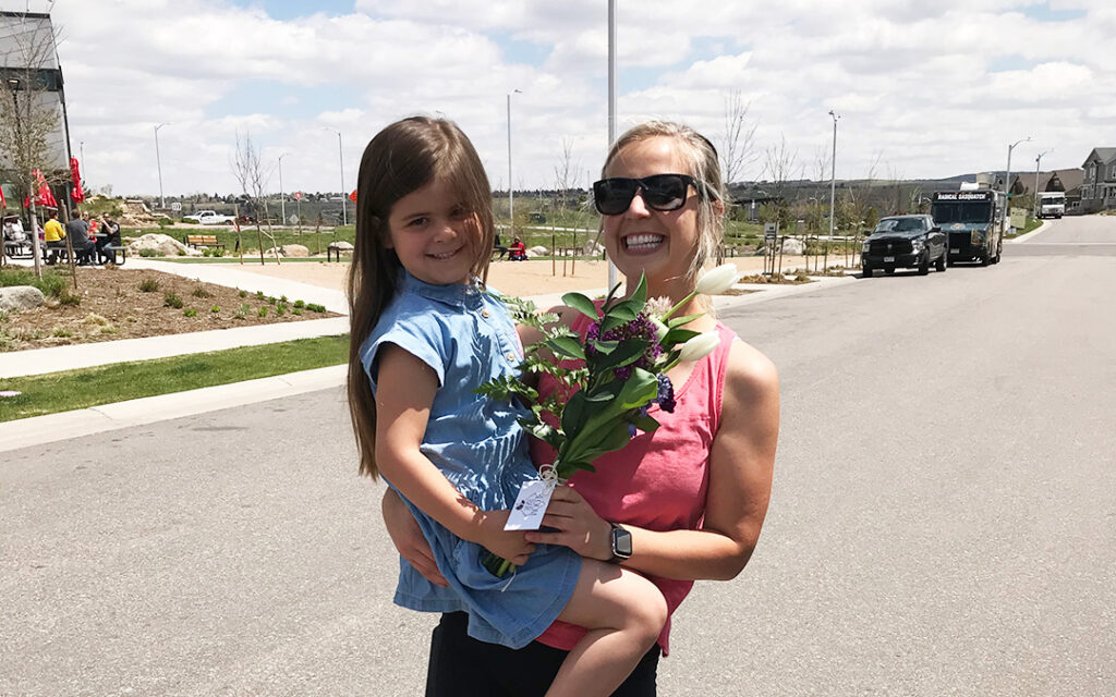 Woman holding a smiling young girl and a bouquet of flowers while standing on a sunny suburban street in Sterling Ranch Colorado, just southwest of Denver.