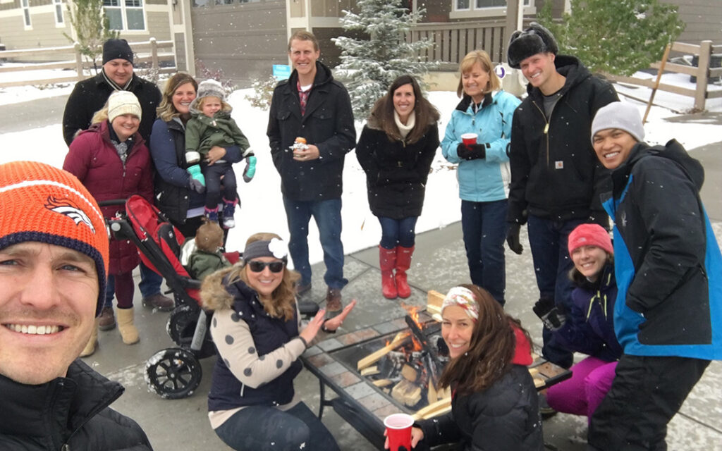 A group of people, including adults and children, gather outdoors around a small firepit on a snowy day at Sterling Ranch Colorado. Some smile at the camera, and others warm their hands by the fire, enjoying a cozy winter moment in the heart of the Denver metro area.