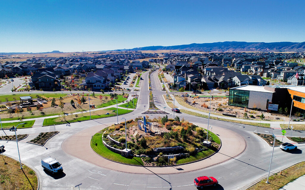 Aerial view of Sterling Ranch, a suburban neighborhood in the Denver metro area, featuring a central roundabout, landscaped greenery, residential houses, and several vehicles on the road under a clear blue sky.