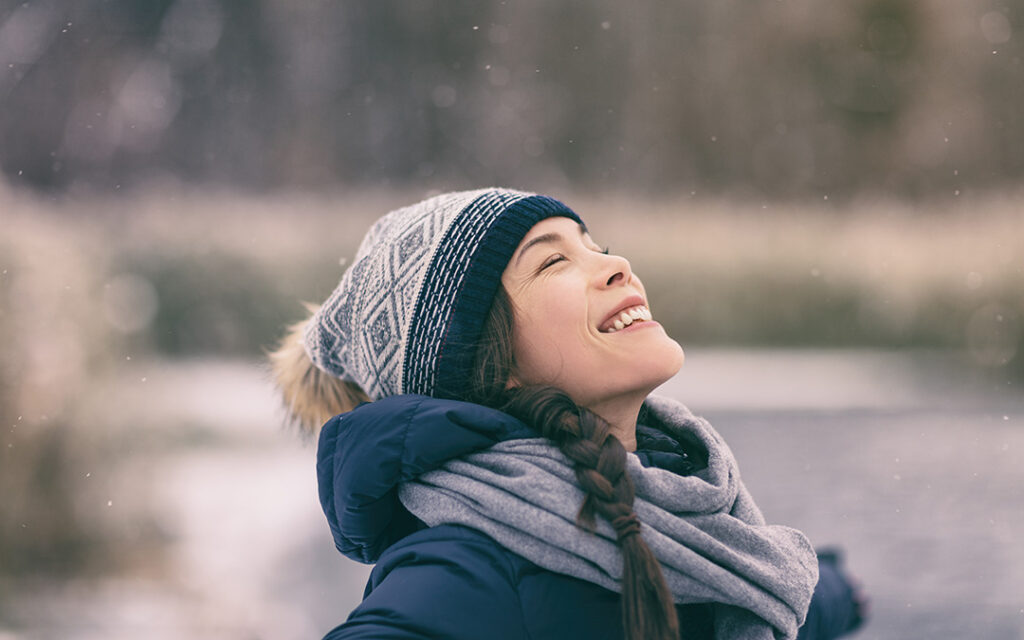 A young person in a winter coat, scarf, and knit hat smiles and looks upward outdoors against the snowy backdrop of Sterling Ranch Colorado.