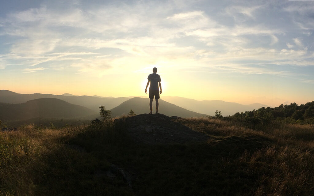 A person stands on a rocky hilltop at sunset, with mountains and a bright sky in the background, capturing the breathtaking views often found southwest of Denver in areas like Sterling Ranch, Colorado.