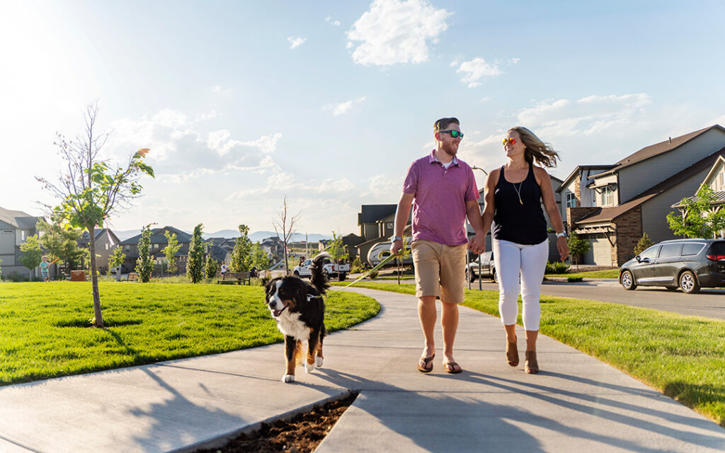 A man and woman walk a dog on a sidewalk in a suburban neighborhood on a sunny day, with houses and parked cars in the background, capturing the vibrant community atmosphere of Sterling Ranch CO, located just southwest of Denver.