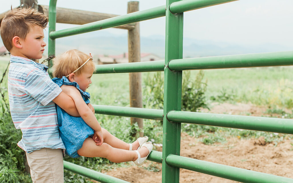 A young boy in summer clothes holds a toddler girl by a green metal fence at Sterling Ranch, with fields and mountains visible in the background.