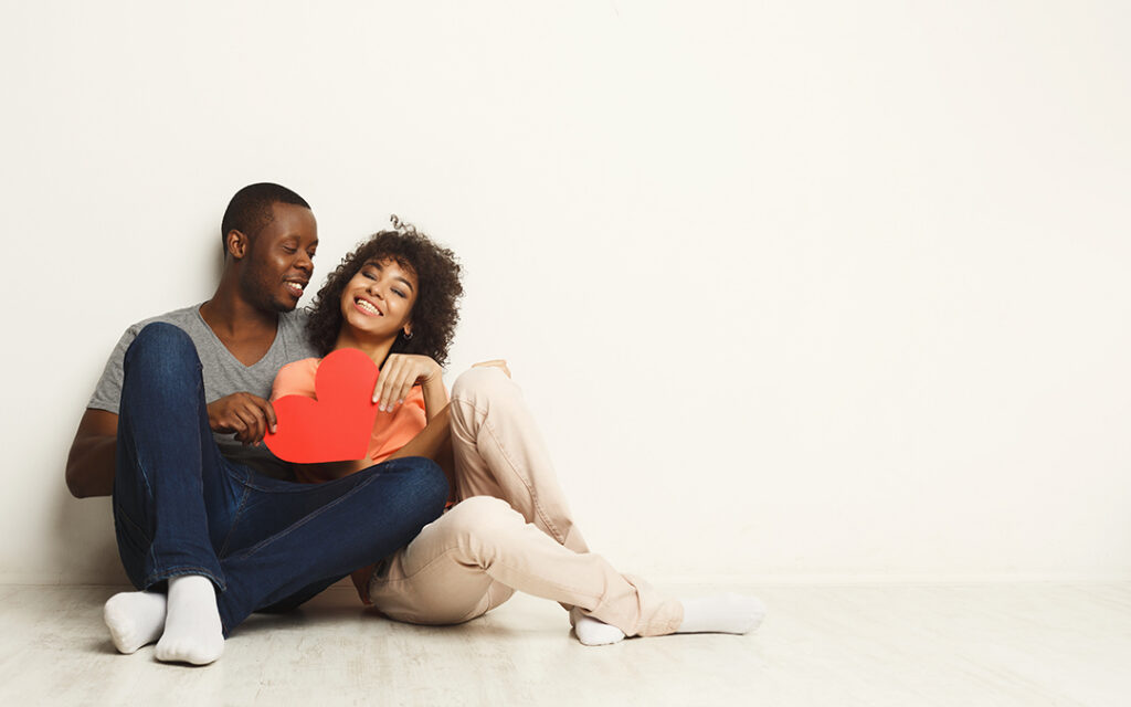 A couple sits on the floor against a white wall, smiling and holding a red paper heart together in their cozy Sterling Ranch CO home, just southwest of Denver.