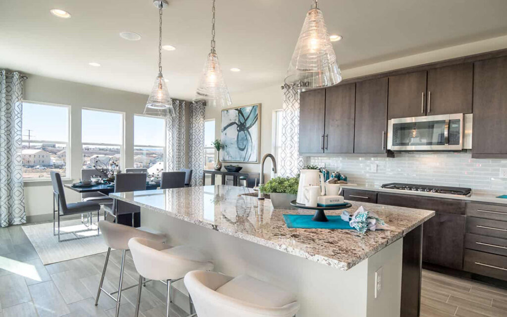 Modern kitchen in Sterling Ranch, Littleton CO, featuring a granite island, gray cabinets, stainless steel appliances, pendant lights, and a dining area with large windows in the background.