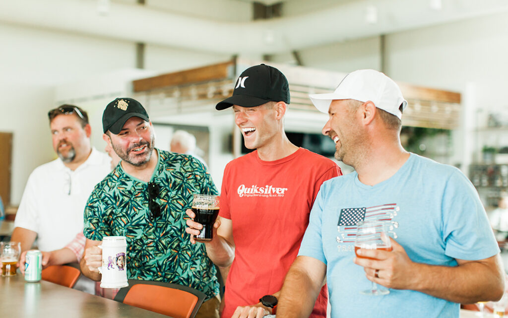 Four men stand at a bar, smiling and laughing while holding drinks, enjoying a lively evening in Sterling Ranch Colorado, just southwest of Denver.