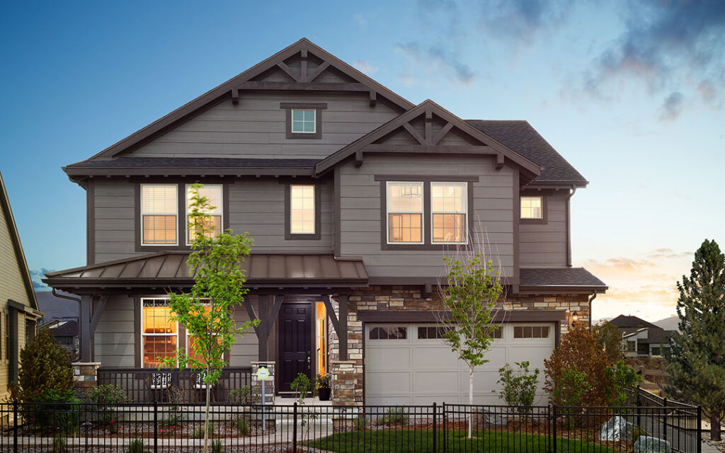 Two-story suburban house with gray siding, stone accents, and a front porch located in Sterling Ranch, southwest of Denver. The house features large windows, a double garage, and a landscaped yard with young trees and shrubs—perfect for enjoying life in the Denver metro area.