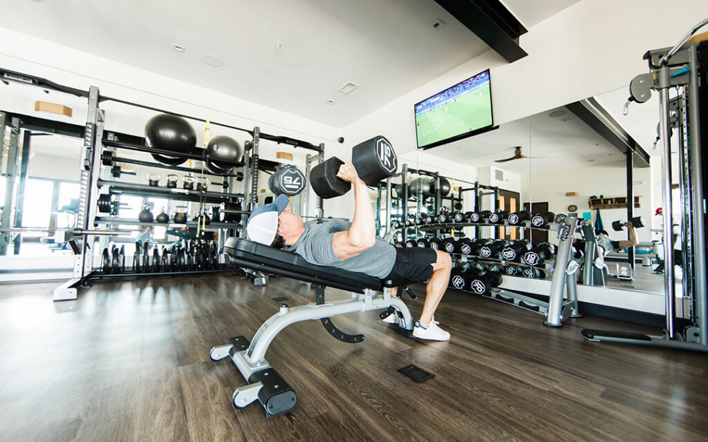 A person in athletic gear does a dumbbell bench press in a modern gym with mirrors, weights, and a TV on the wall, showcasing the high-quality fitness facilities available in Sterling Ranch Colorado.
