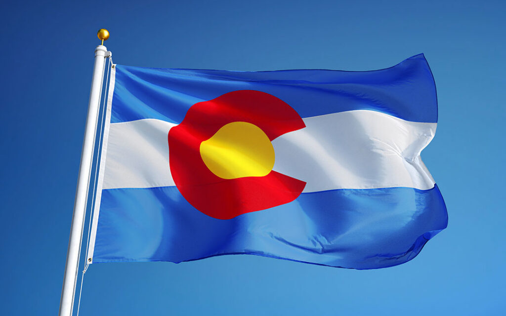 The Colorado state flag features blue and white stripes with a circular red "C" surrounding a golden disk, flying on a flagpole against a clear blue sky in the vibrant community of Sterling Ranch CO.