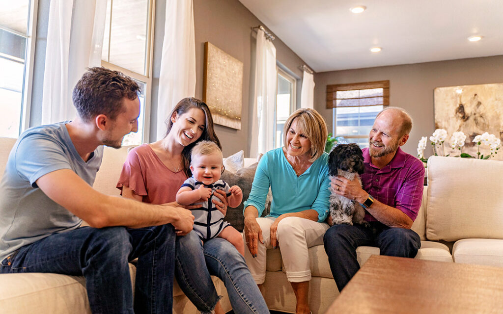 Three adults, a baby, and an older couple with a small dog sit together on a living room couch, smiling and talking in a bright, cozy home in Sterling Ranch, just southwest of Denver.