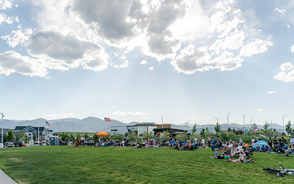 People sit on a grassy field in a park on a sunny day, with flags, mountains, and buildings in the background—capturing the welcoming community spirit that defines Sterling Ranch CO in the greater Denver metro area.