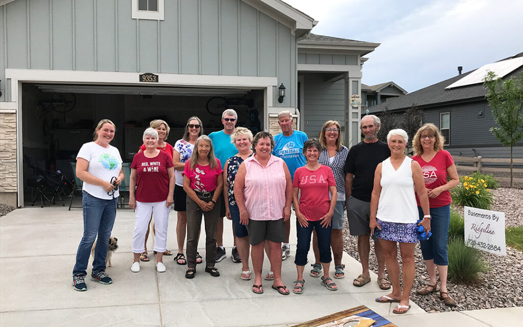 A group of people stand and smile in front of a house on a driveway, with some wearing patriotic-themed shirts; the house number 3633 is visible above the garage at Sterling Ranch Colorado, a welcoming neighborhood in the Denver metro area near Littleton CO.