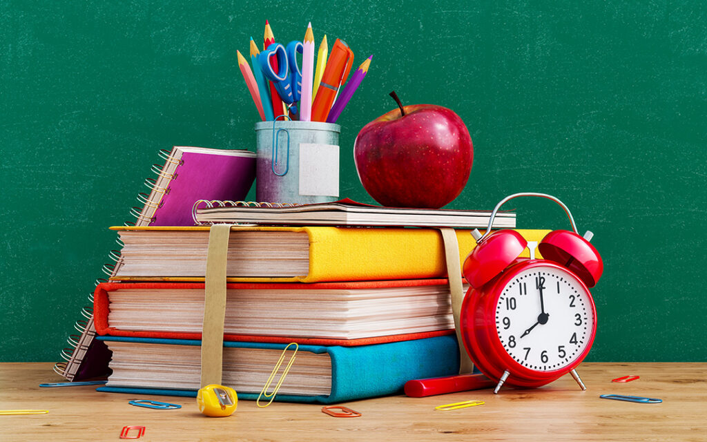 Stack of books with notebooks, a red apple, a cup of colored pencils, paper clips, a pencil sharpener, and a red alarm clock arranged on a desk in front of a green chalkboard—perfect for inspiring young minds whether you're in the heart of the Denver metro or exploring new learning spaces in Sterling Ranch CO.