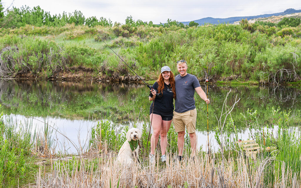 Two people stand by a pond holding fishing rods, smiling at the camera, with a white dog sitting in front of them among grass and reeds in Sterling Ranch CO, southwest of Denver.