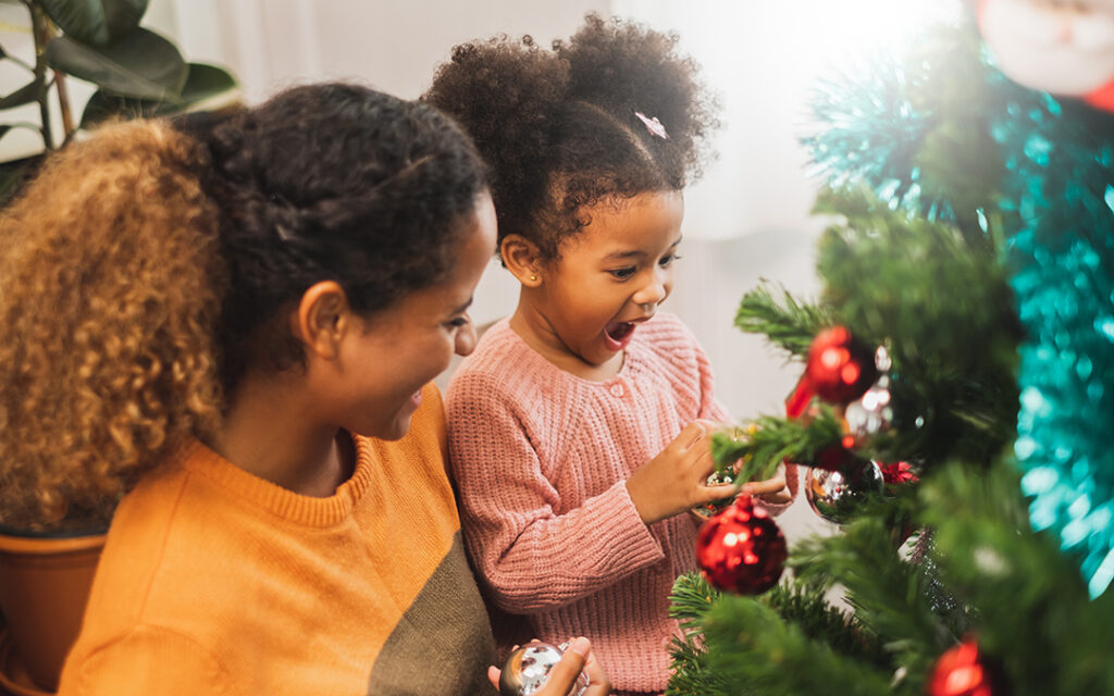 An adult and a child decorate a Christmas tree with ornaments in their cozy home southwest of Denver. The child looks excited while placing a decoration on the tree, bringing festive cheer to this charming corner of the Denver metro area.