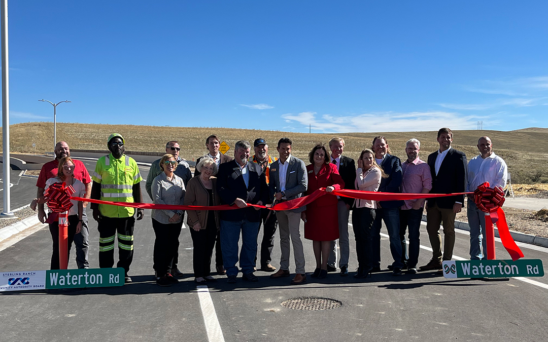A group of people stands behind a red ribbon at a road opening ceremony, holding Waterton Rd signs, with open land and blue sky in the background at Sterling Ranch Colorado, just southwest of Denver in the Denver metro area.