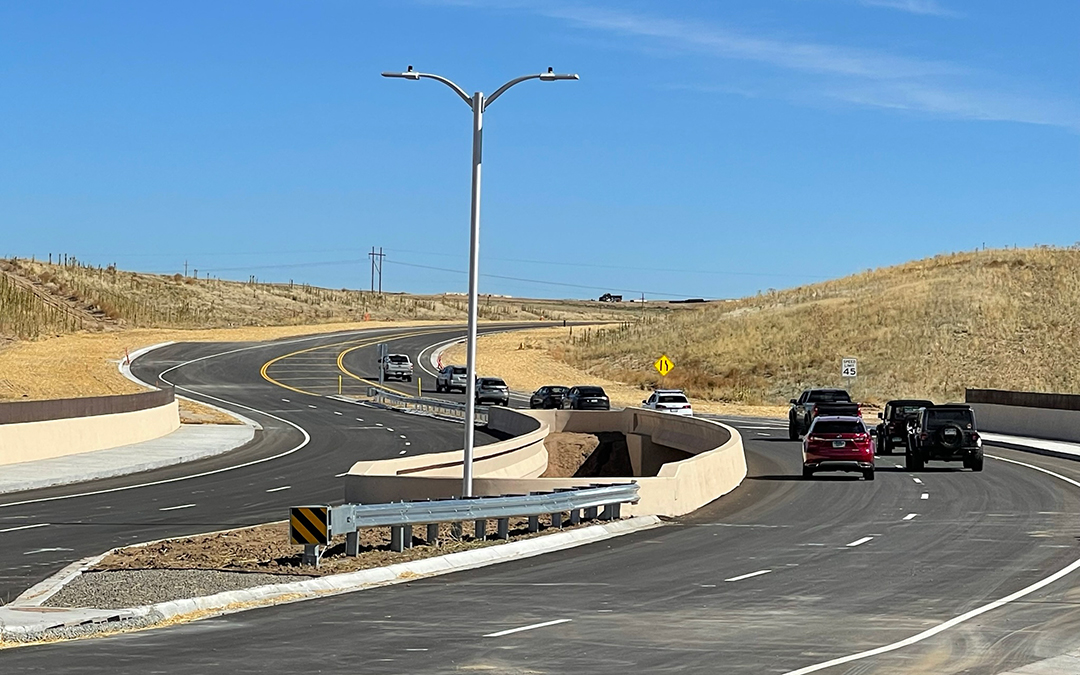 Cars travel on a newly paved divided road with light traffic, surrounded by the dry, grassy hills of Sterling Ranch Colorado under a clear blue sky, just outside the Denver metro area.